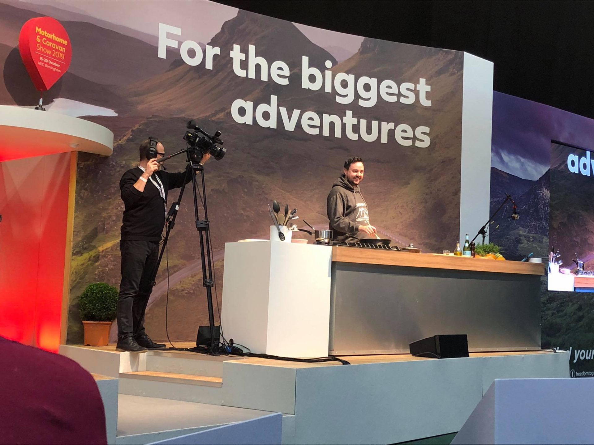 Michael Basco giving a live open-fire cooking demonstration on stage at the Motorhome and Caravan Show, filmed by a TV cameraman